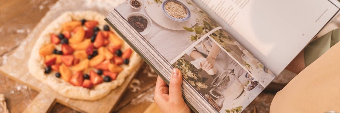 A person reads a cookbook while preparing a fruit tart on a wooden table.