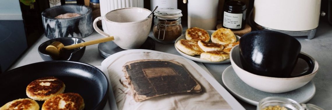 One portion of homemade cheese pancakes in frying pan and another on plate locating near opened stylish magazine among kitchen utensils and appliances in bright kitchen at home